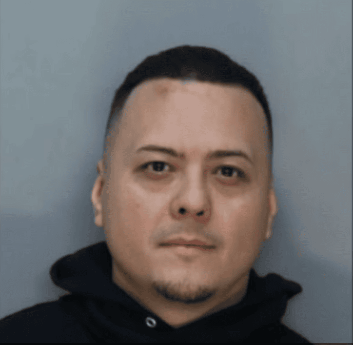 A man with short dark hair and a thin beard is wearing a black hoodie, embodying the intensity of a law enforcer. The backdrop is plain grey, and his neutral expression hints at the determination of someone who might be involved in arresting those smuggling narcotics.